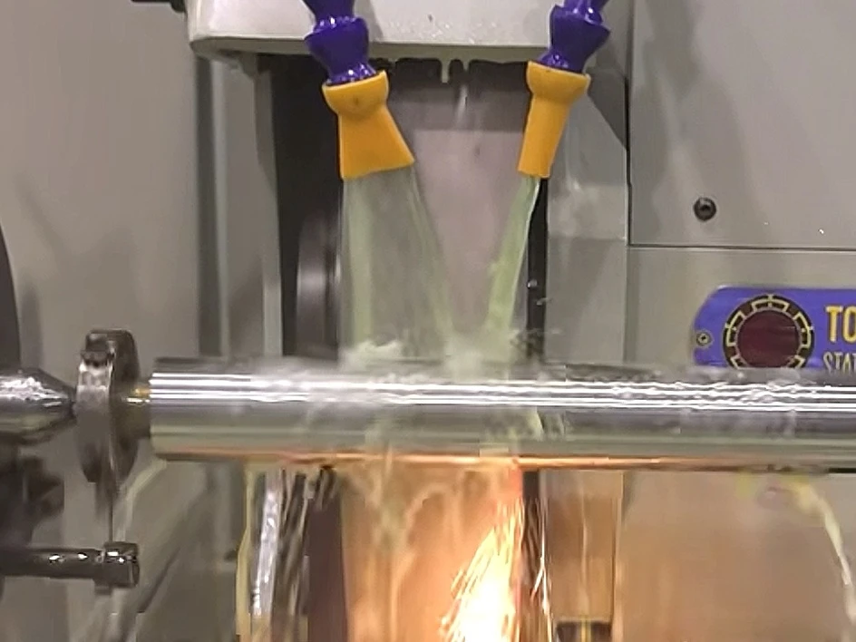 A cylindrical metal shaft being ground with coolant at the grinding wheel